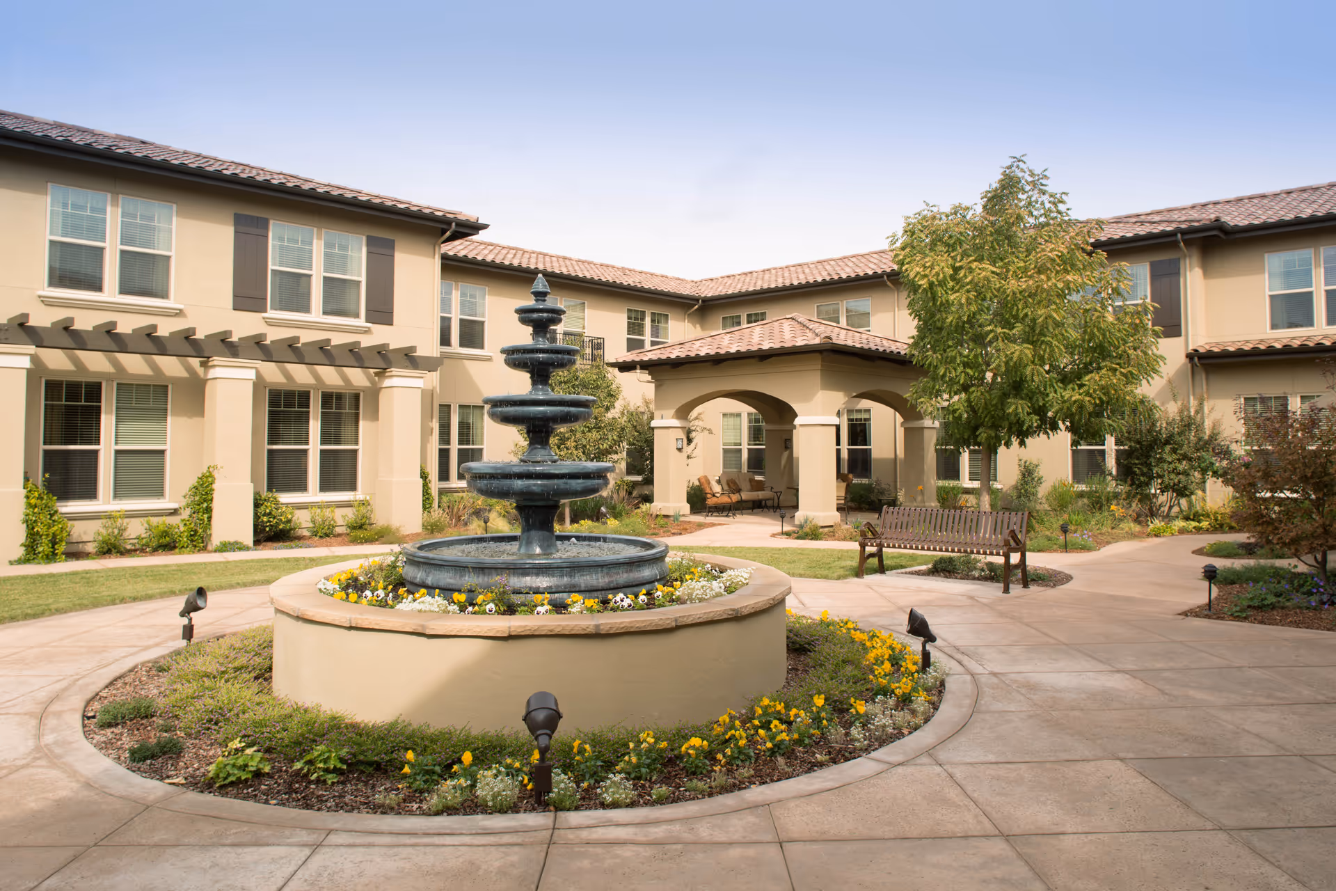 Outdoor courtyard area of a senior living facility with a multi-tiered water fountain surrounded by flowers in the center. The courtyard has paved walkways, benches, trees, and shrubs, with a two-story beige building with multiple windows and a covered seating area in the background.