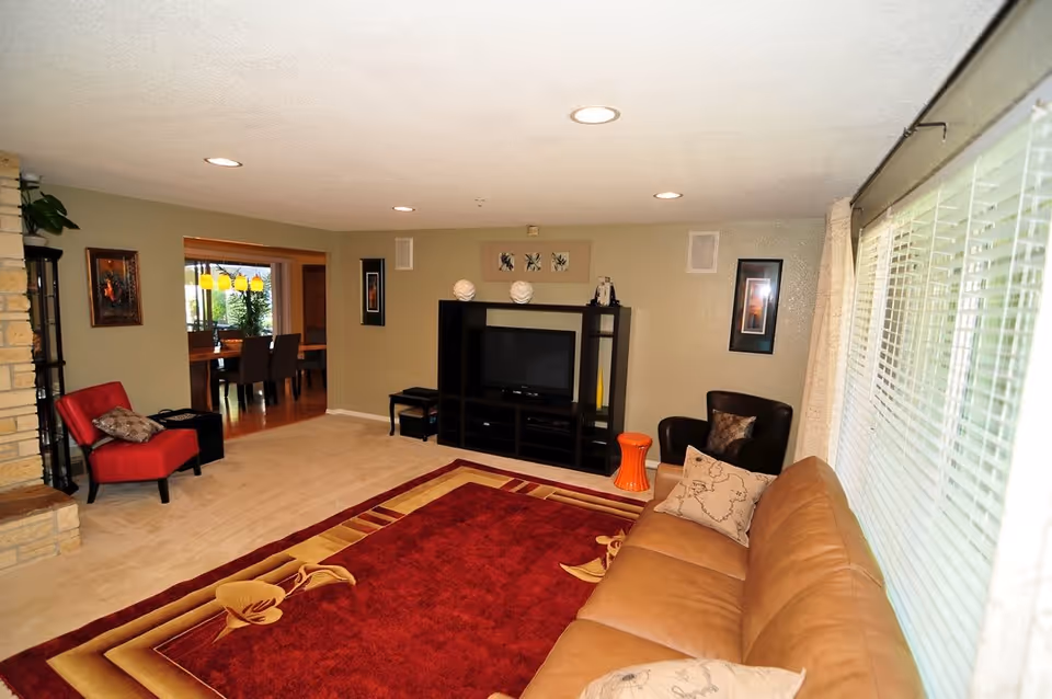 A cozy living room with a large window covered by white blinds on the right side. The room features a tan leather sofa with decorative pillows, a red and beige patterned area rug, a black entertainment center with a TV, and two chairs—one red and one dark brown. The walls are painted light beige, and there are framed artworks on the walls. In the background, a dining area with a wooden table and chairs is visible through an open doorway.