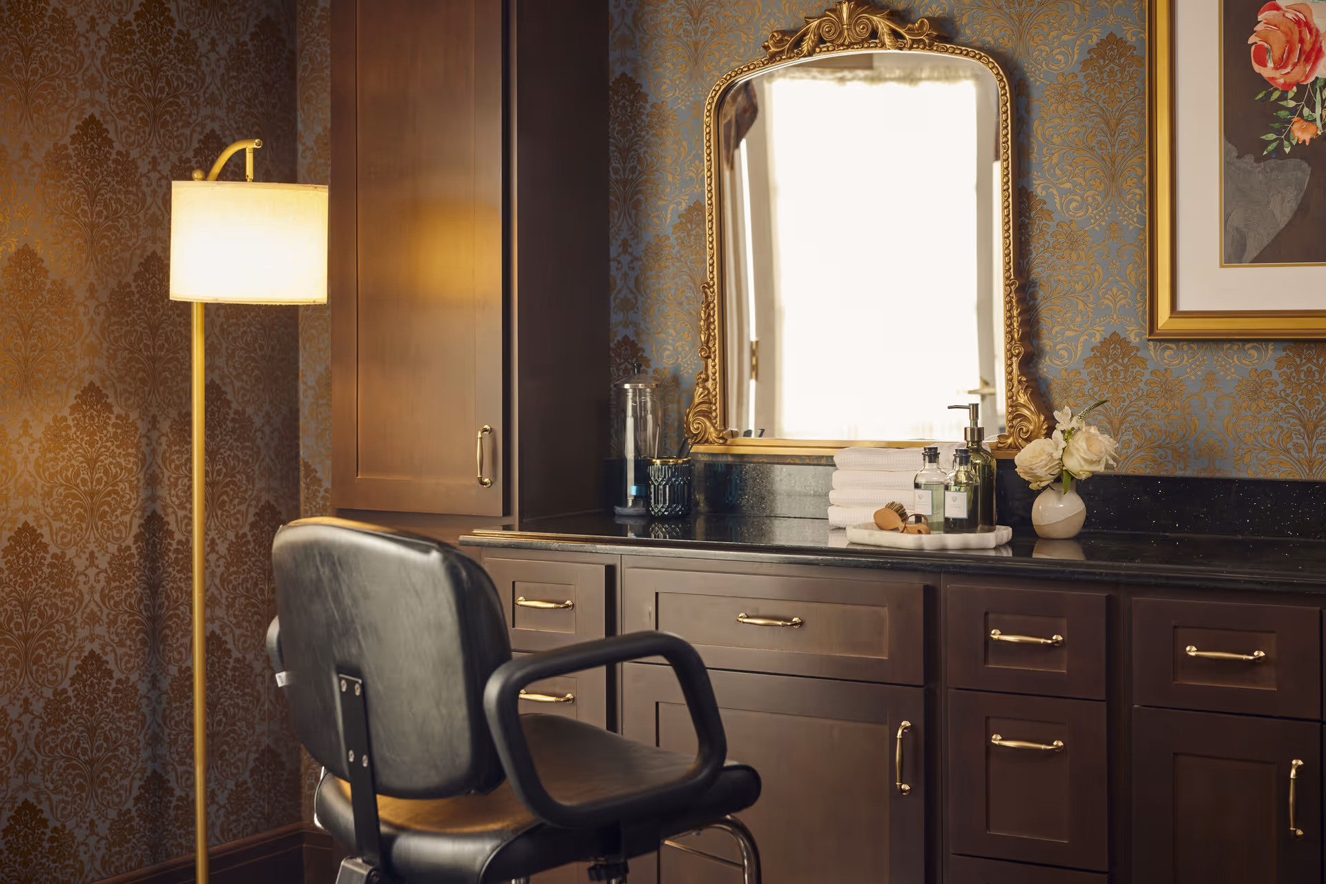 A vintage-style vanity area with a large ornate gold-framed mirror, dark wood cabinets with gold handles, a black cushioned chair, a standing lamp with a beige shade, and decorative items including folded white towels, bottles, a brush, and a small vase with white roses. The wall has a patterned gold and blue wallpaper and a framed floral painting.