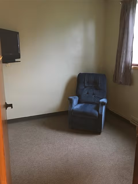 A small, sparsely furnished room with beige walls and carpeted floor. There is a single blue upholstered armchair positioned near the corner, a window with gray curtains on the right wall, and a wall-mounted flat-screen TV on the left wall.
