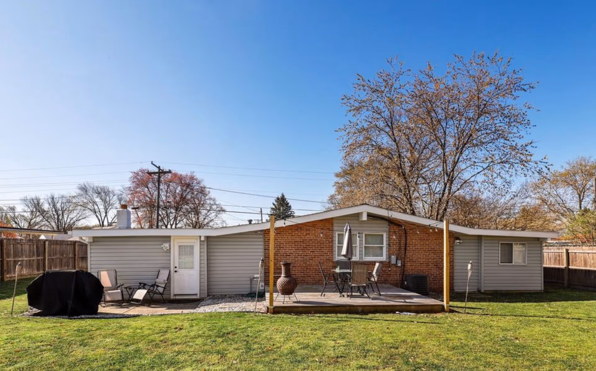 Single-story brick and siding building with a small wooden patio, outdoor furniture, a grill, and a grassy fenced backyard under a clear blue sky.