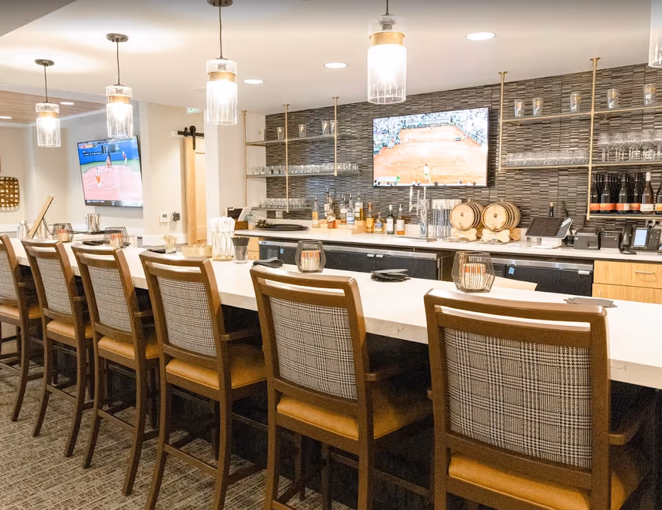 Interior view of a modern bar area with a long white countertop and eight wooden chairs with plaid backs and leather seats. Behind the bar are shelves with glasses, bottles, and two small barrels. Two flat-screen TVs are mounted on the walls, one showing a tennis match and the other showing a softball game. Pendant lights hang from the ceiling above the bar.