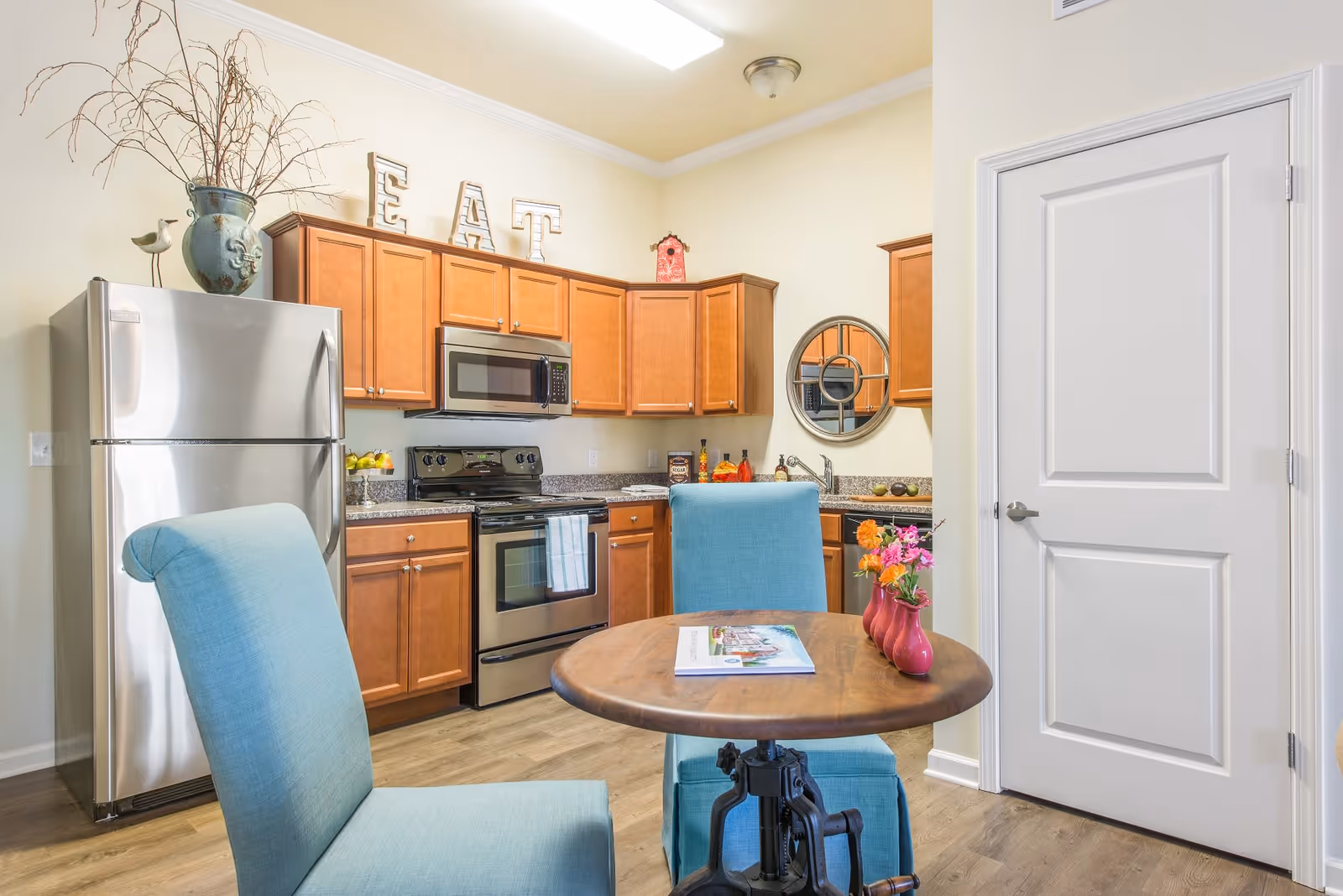 A bright kitchen area with wooden cabinets, stainless steel refrigerator, stove, and microwave. A round wooden table with two blue upholstered chairs is in the foreground, with a small vase of flowers and a magazine on the table. Decorative letters spelling 'EAT' are displayed on top of the cabinets, and a round mirror hangs on the wall above the sink.