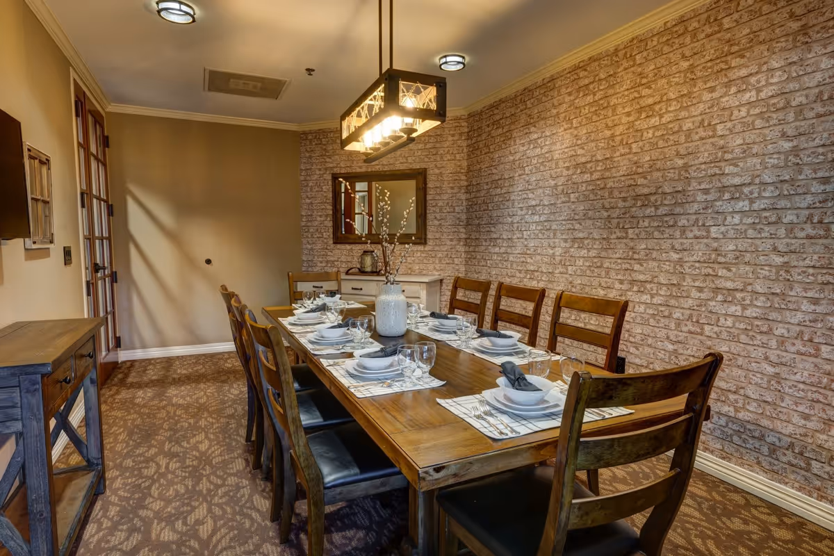 A dining room with a long wooden table set for eight people. The table has white plates, bowls, glasses, silverware, and gray napkins on plaid placemats. A white vase with decorative branches is centered on the table. The room features a brick accent wall, a mirror, a wooden sideboard, and a patterned carpet. There is a modern rectangular chandelier hanging above the table and two ceiling lights.