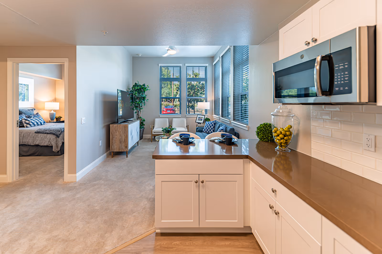 View from a kitchen area into a living room and bedroom in a senior living facility. The kitchen has white cabinets, a brown countertop, and a microwave. The living room features a TV on a wooden stand, two chairs, a blue couch with pillows, a small table with a lamp, and large windows with blinds letting in natural light. The bedroom visible through an open door has a bed with gray bedding and a bedside table with a lamp.