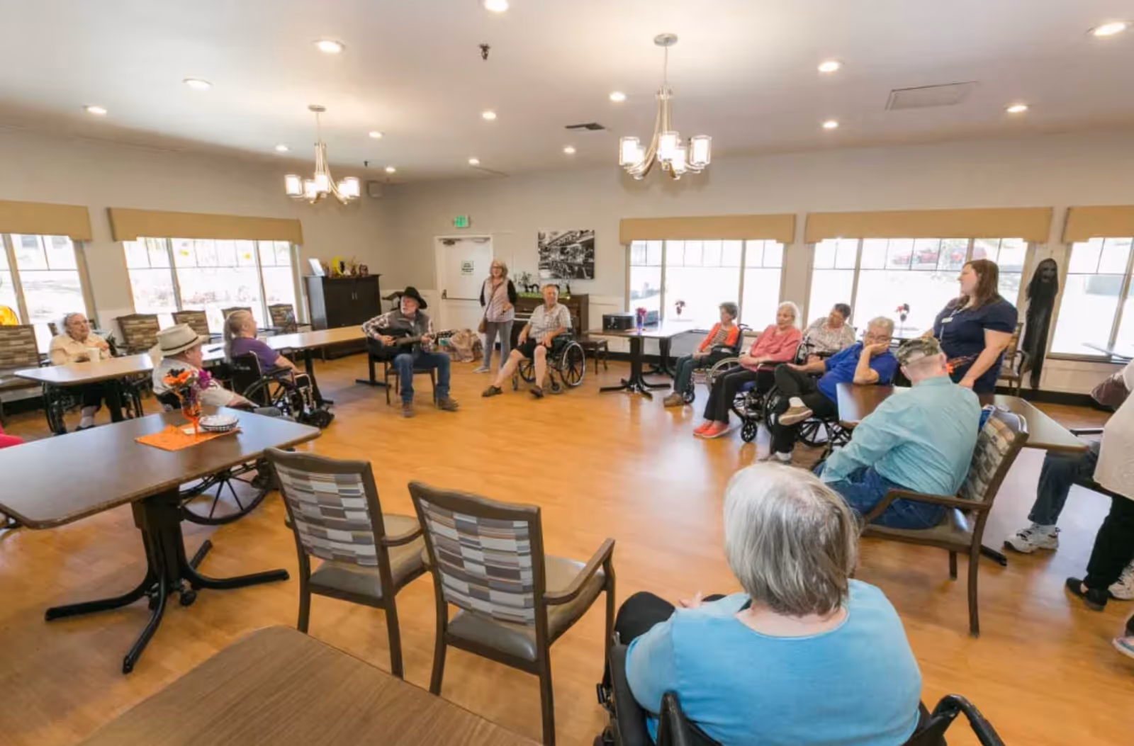 Elderly residents sitting around tables in a nursing home common room while a musician plays guitar.
