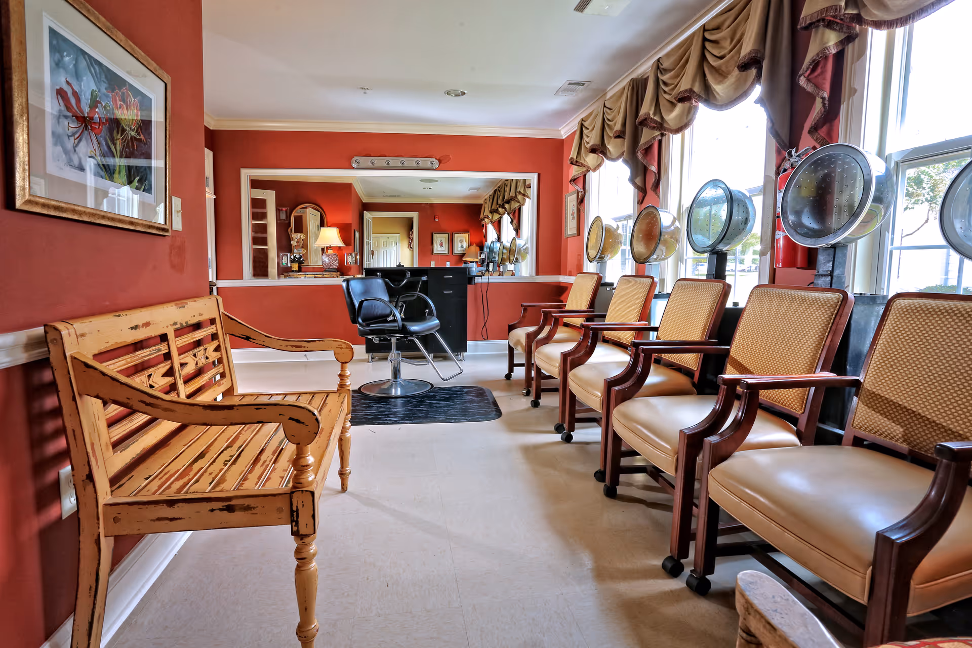 A bright salon waiting area with a row of vintage hooded hairdryers and cushioned armchairs along sunlit windows, plus a wooden bench and styling chair near a mirror.