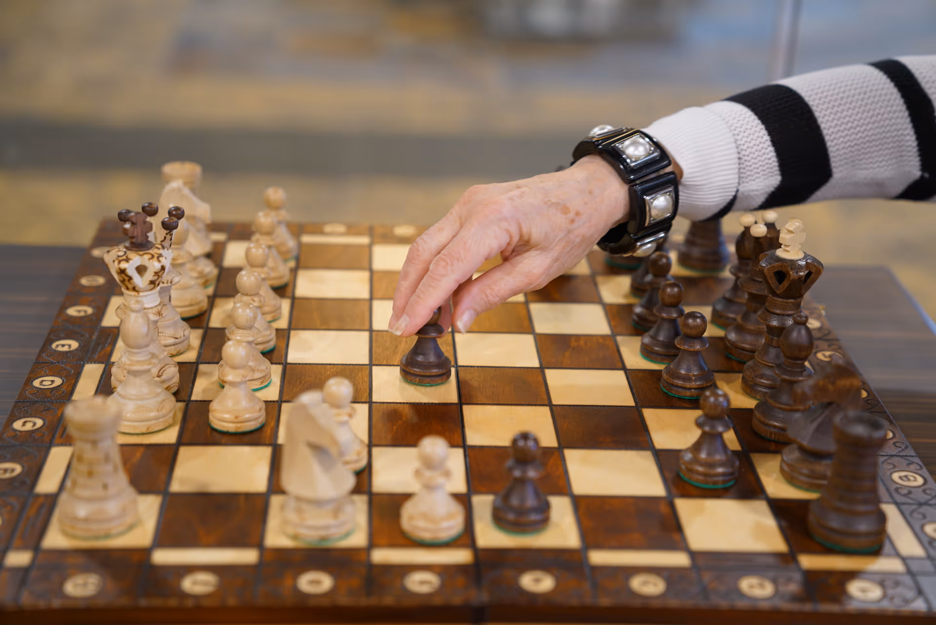 A close-up of an elderly person's hand moving a dark chess piece on a wooden chessboard with both light and dark pieces arranged for a game. The person is wearing a black and white striped sweater and a black bracelet with large square decorations.