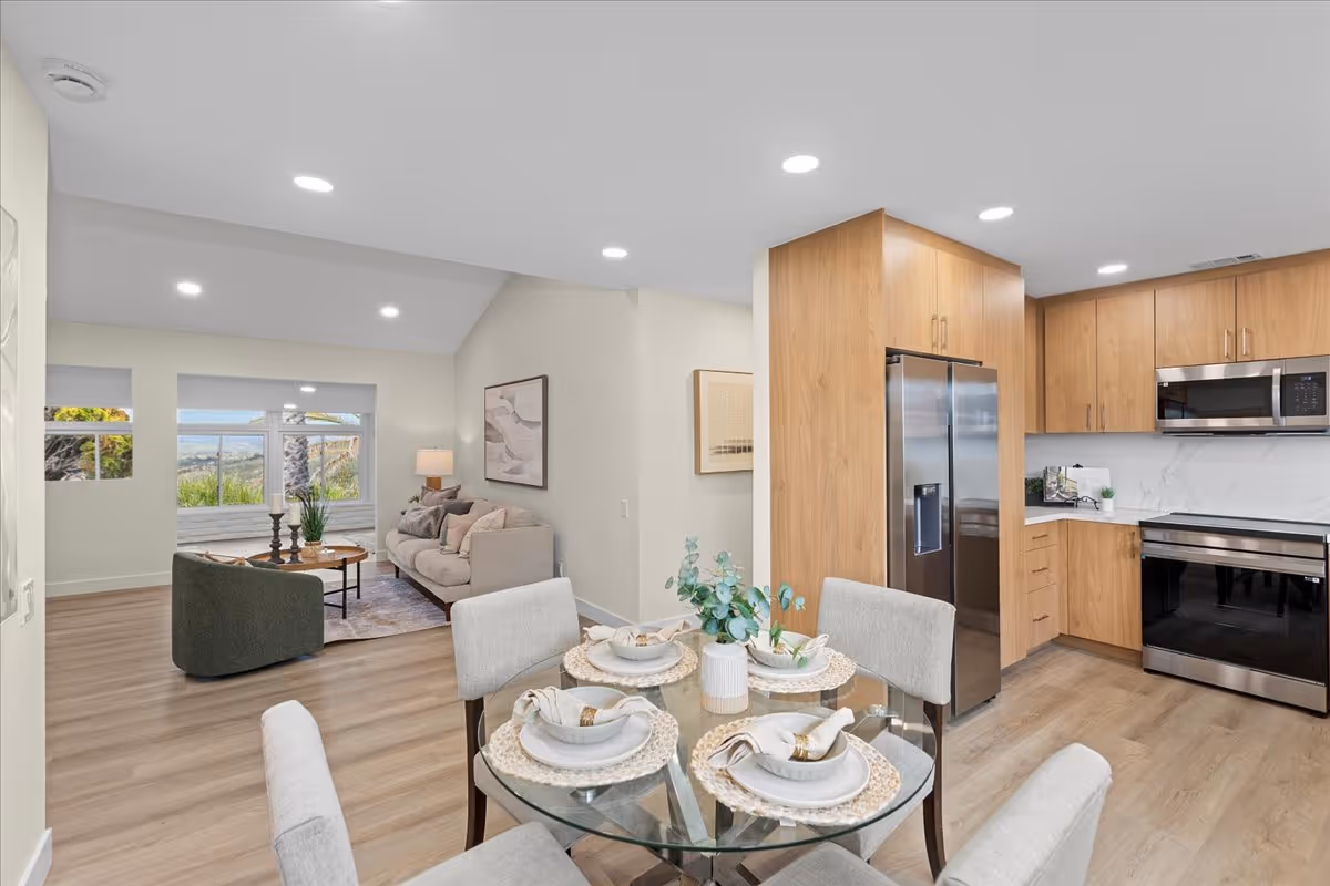 Open-plan interior with a dining table in the foreground, a seating area by a large window, and a modern kitchen with wood cabinets.