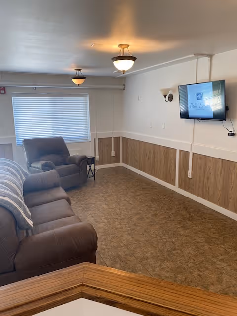 A cozy living room area with brown cushioned sofas and armchairs arranged along the walls. The room has a window with closed blinds, two ceiling light fixtures, and a wall-mounted TV displaying the Palm Garden logo. The walls are white with wood paneling on the lower half, and the floor is covered with brown patterned linoleum.
