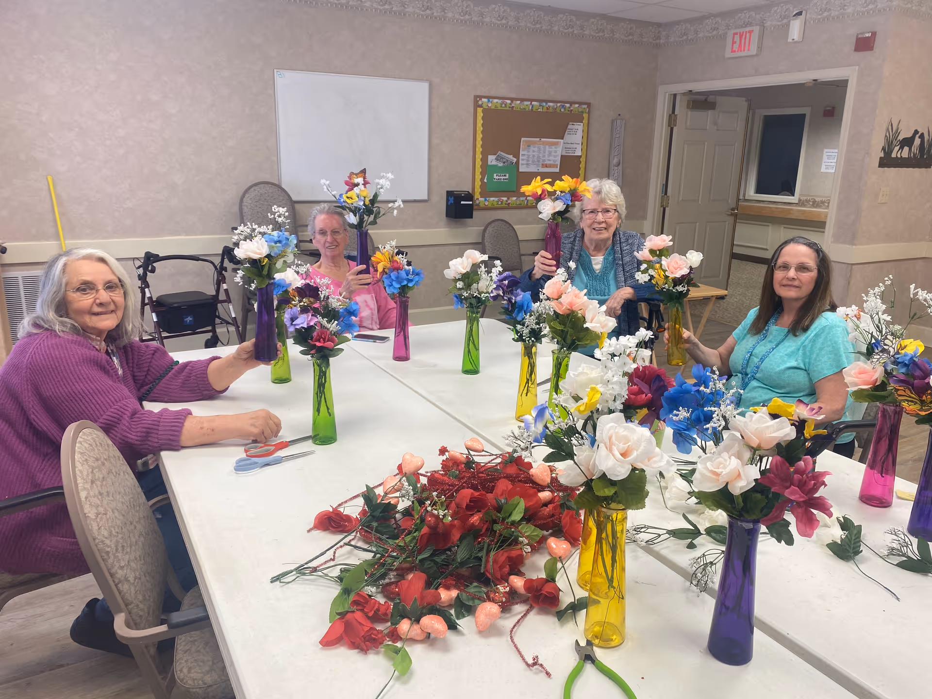 Four elderly women sitting around a table in a senior living facility, each holding colorful flower arrangements in tall vases. The table is covered with various artificial flowers and floral supplies. The room has beige walls, a bulletin board, and an exit sign above a door in the background.
