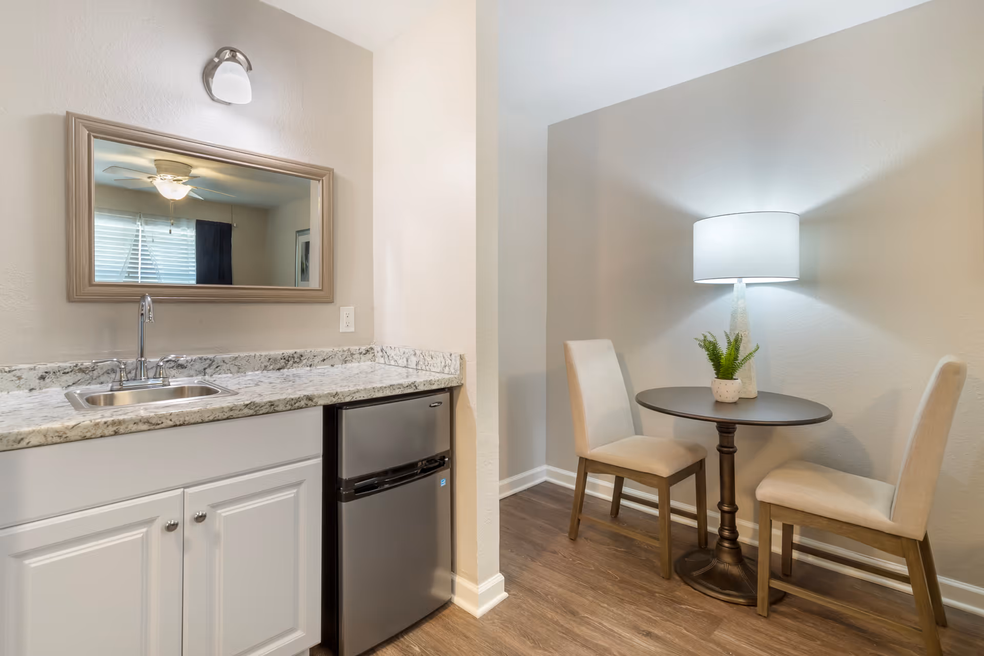A small kitchenette area with a granite countertop, a stainless steel sink, and a mini refrigerator. Adjacent to the kitchenette is a small round table with two beige upholstered chairs. A table lamp with a white shade and a small potted plant are on the table. The room has light-colored walls and wood flooring.