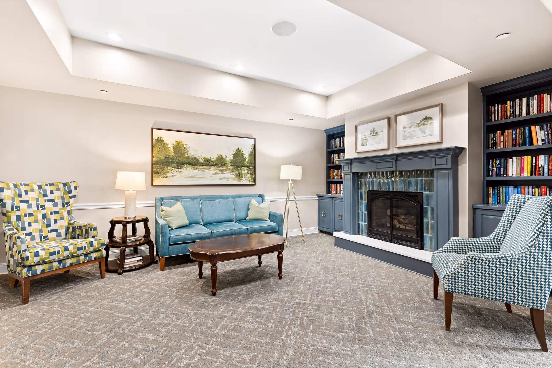 A cozy living room with a blue sofa, two patterned armchairs, a wooden coffee table, and a side table with a lamp. The room features a fireplace with blue tiles and built-in bookshelves filled with books on either side. Above the fireplace are two framed landscape paintings. The walls are light-colored, and the carpet has a subtle pattern.
