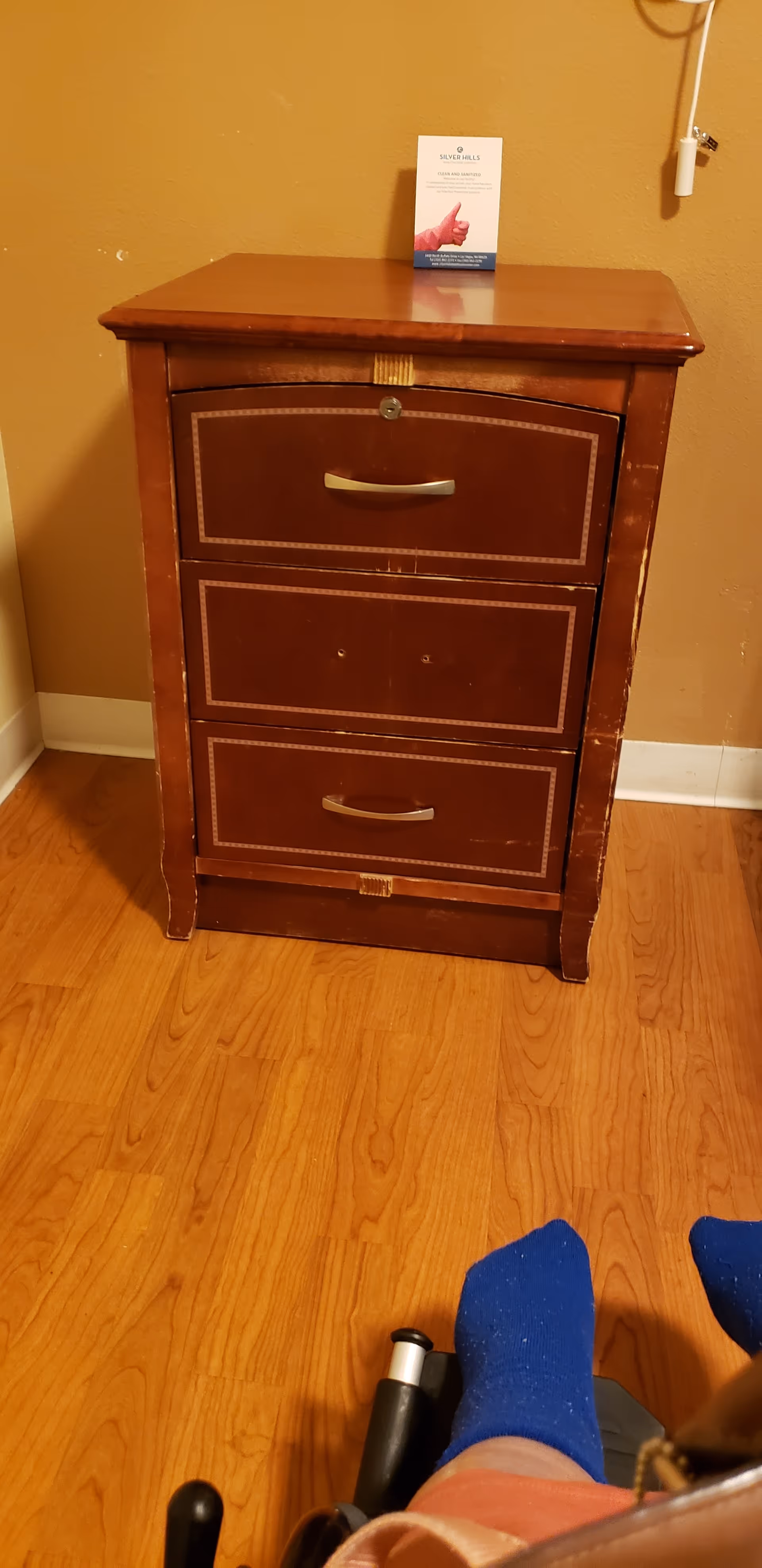 A wooden three-drawer dresser against a tan wall with a card on top.