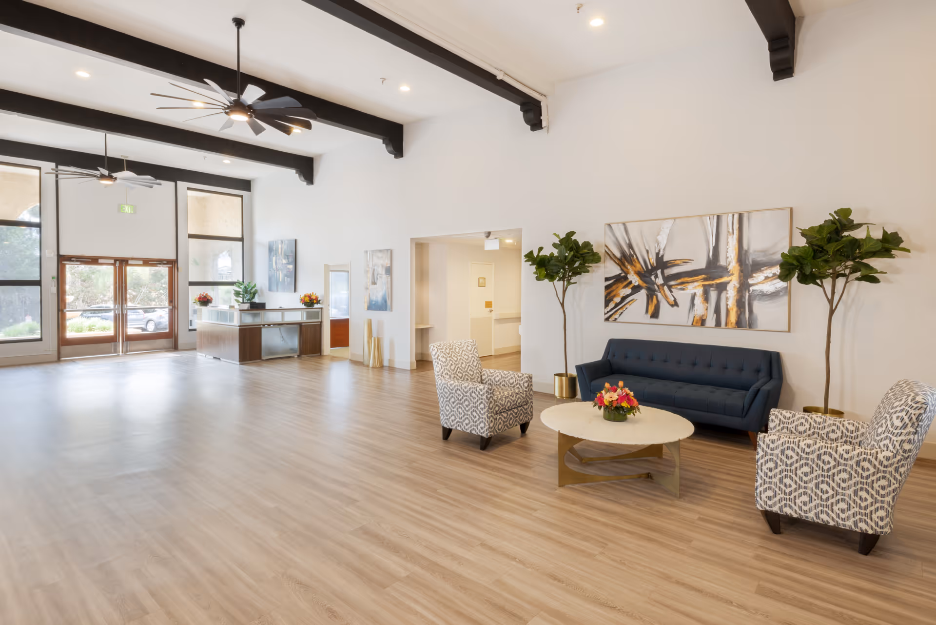 A spacious and bright healthcare center lobby with light wood flooring, high ceilings with exposed dark beams, and large windows letting in natural light. The seating area includes a dark blue sofa, two patterned armchairs, and a round coffee table with a flower arrangement. There are two tall potted plants flanking an abstract painting on the wall. A reception desk is visible near the entrance with floral decorations.