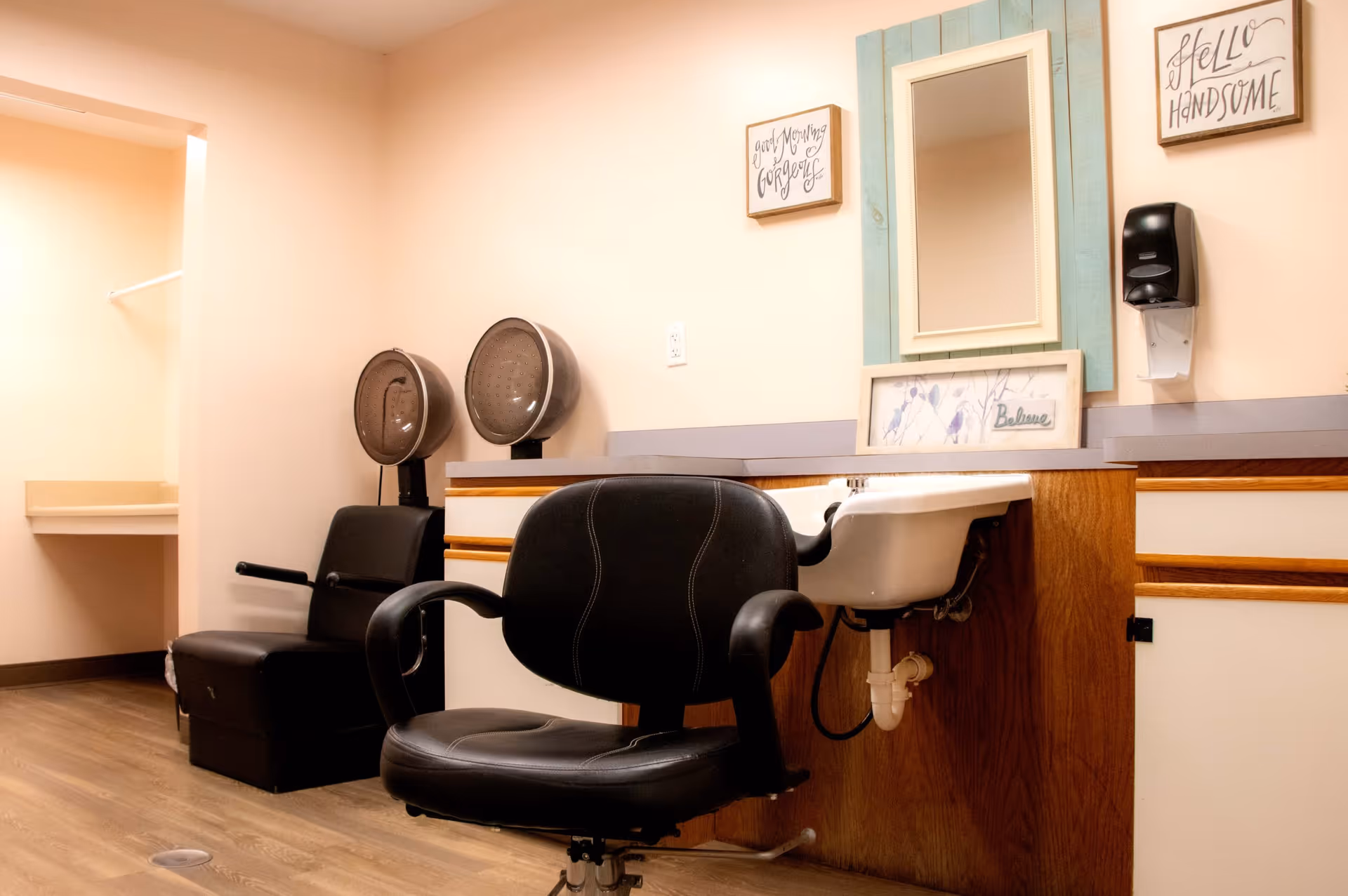 Salon station inside a senior living facility with a styling chair, shampoo sink, hooded dryers, mirror, and countertop.