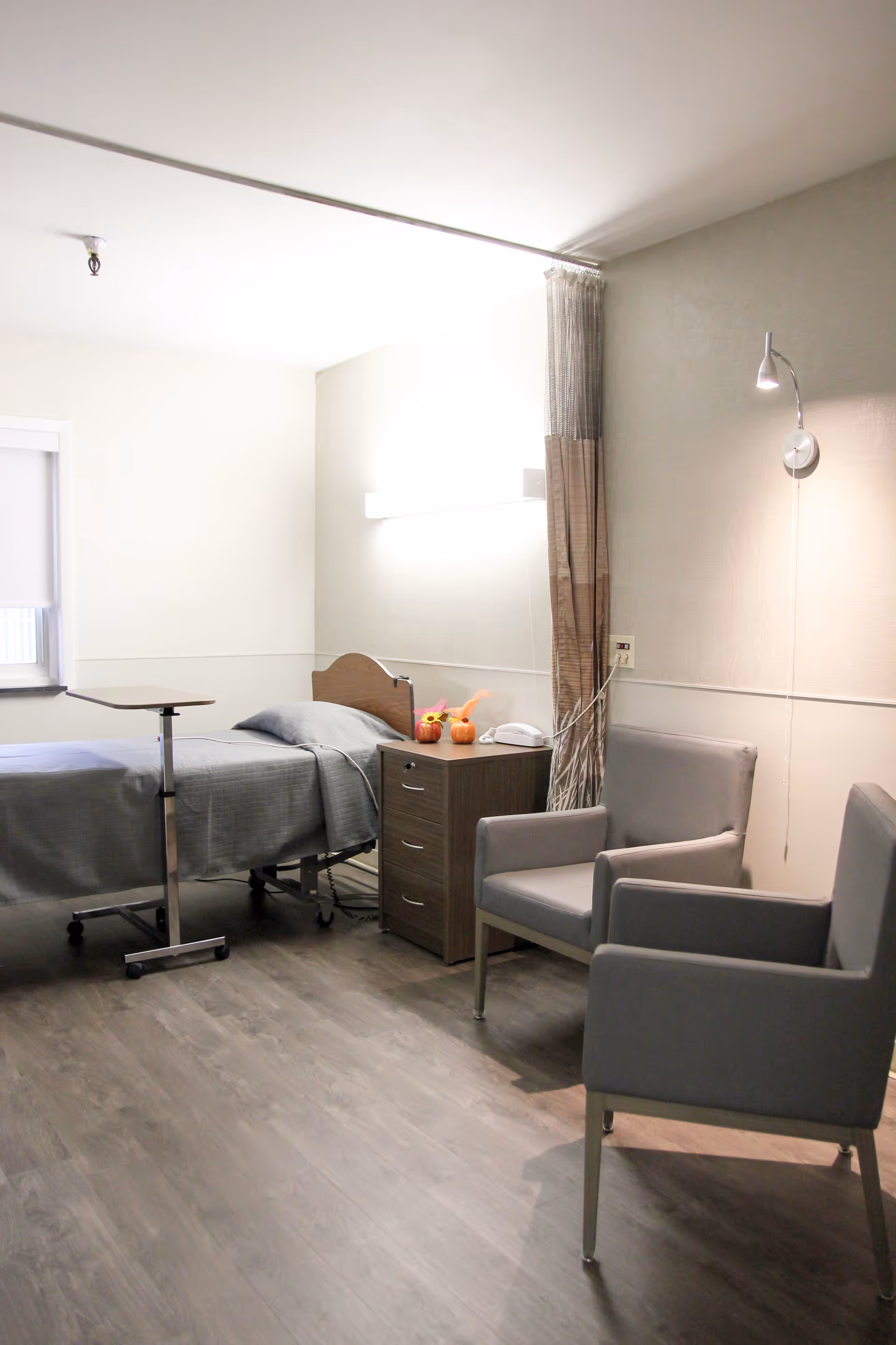 A simple, clean patient room with a single bed covered in gray bedding, a small bedside table with a telephone and decorative items, two gray armchairs, a wall-mounted reading light, and a window with a roller shade partially drawn.