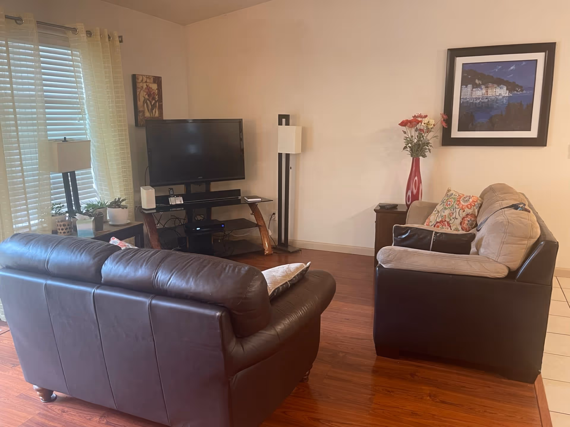 A cozy living room with two sofas, one dark brown leather and the other beige with dark brown sides, arranged around a TV on a stand. There is a floor lamp next to the TV, a side table with a red vase holding flowers, and a framed picture on the wall. The room has wooden flooring and a window with light-colored curtains and blinds.