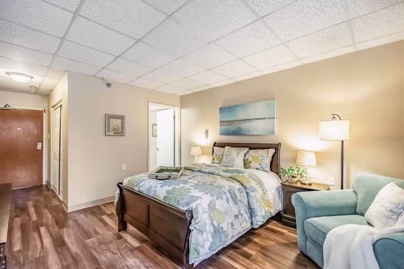 A cozy furnished bedroom with a floral-patterned bed, wooden bedframe, nightstands and lamps, a green armchair, and wood-look flooring.