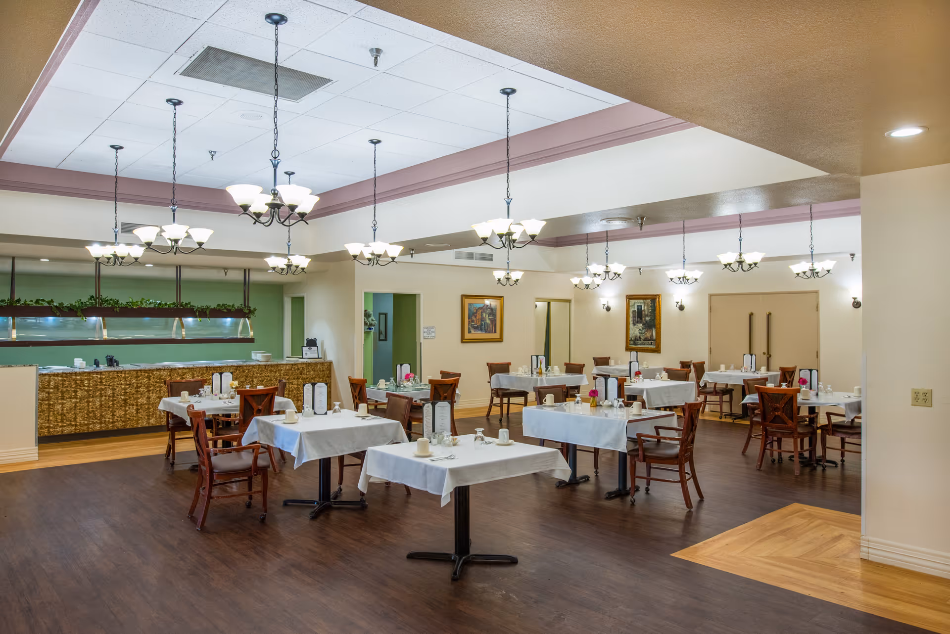 A spacious dining room with multiple tables covered in white tablecloths, each set with cups, glasses, and menus. The room features wooden chairs, several chandeliers hanging from the ceiling, framed artwork on the walls, and a serving counter with a decorative front and greenery above it.