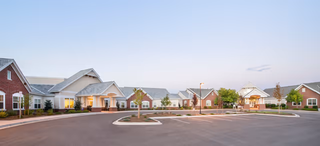 Exterior view of Brookdale Highlands Ranch senior living facility at dusk, showing a large parking area in front of a single-story building with multiple entrances, brick and siding facade, and well-maintained landscaping.