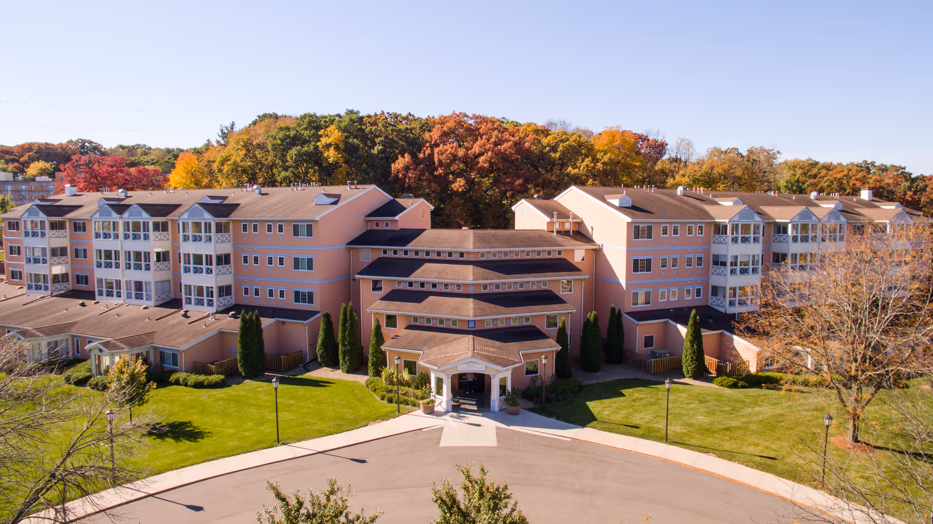 Aerial view of a large, multi-story senior living facility building with peach-colored exterior walls and multiple windows. The building is surrounded by well-maintained green lawns, trees with autumn foliage, and a circular driveway at the entrance. The background features a forested area with trees in fall colors under a clear blue sky.