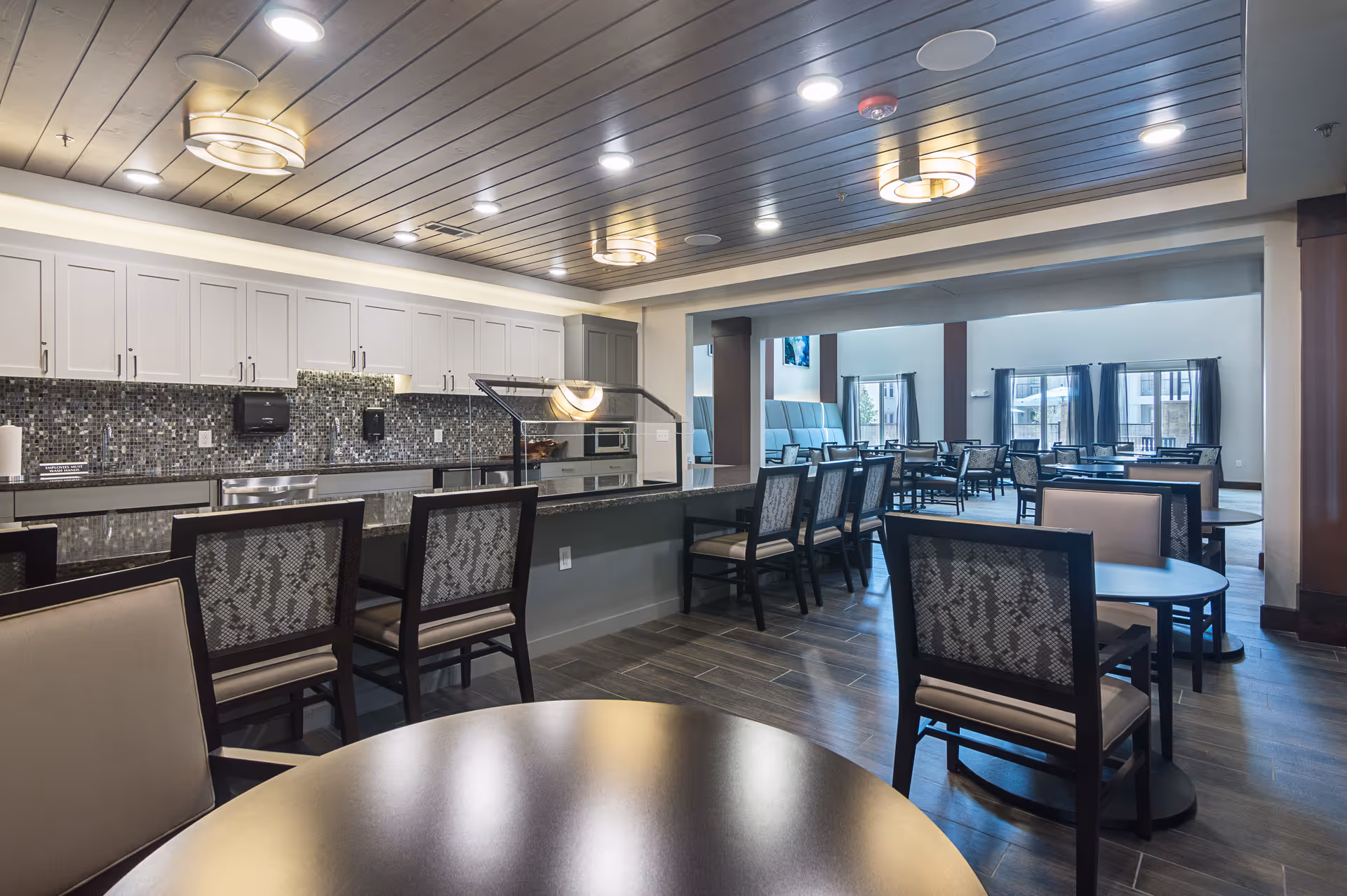 A spacious dining area in a senior living facility with multiple tables and chairs arranged neatly. The room features a serving counter with a mosaic tile backsplash, white cabinets, and modern ceiling lights. Large windows in the background allow natural light to fill the space.