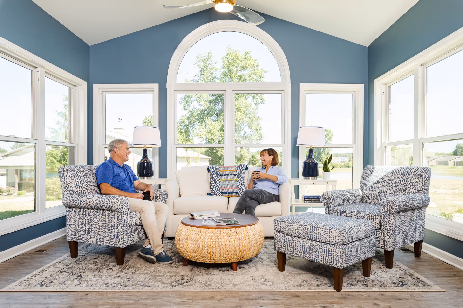 A bright living room with large windows and blue walls, featuring a beige sofa with a colorful pillow, two patterned armchairs, and a matching ottoman. A round wicker coffee table with a wooden top sits on a patterned rug. Two table lamps with dark blue bases and white shades are on white side tables. An older man and woman are sitting and smiling while holding coffee mugs.