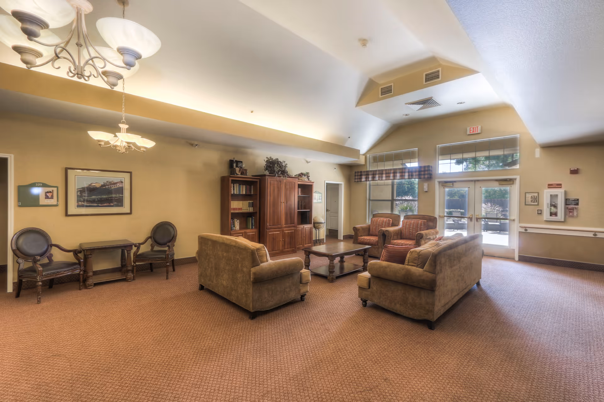 A spacious senior living facility common area with beige walls and carpet. The room features two brown sofas and two armchairs arranged around a wooden coffee table. There is a wooden cabinet with books and decorative items against the wall. Large windows and glass doors let in natural light, and there are ceiling lights and a chandelier providing additional lighting. A small table with two chairs is positioned near the wall with framed artwork above it.