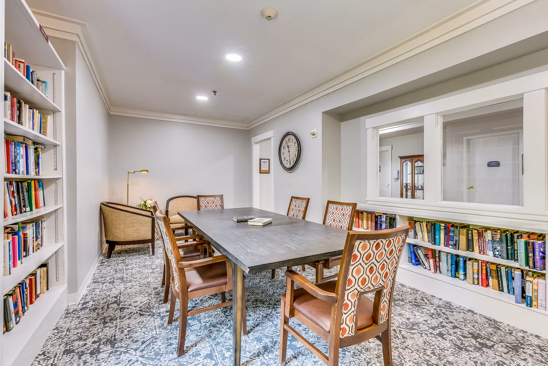 A cozy library-style meeting room with a rectangular table surrounded by chairs, bookshelves, and a reading nook.