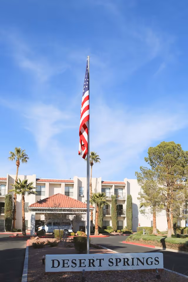 Exterior view of Desert Springs Senior Living facility with a clear blue sky, an American flag on a flagpole in the foreground, palm trees, and a three-story building with balconies in the background.