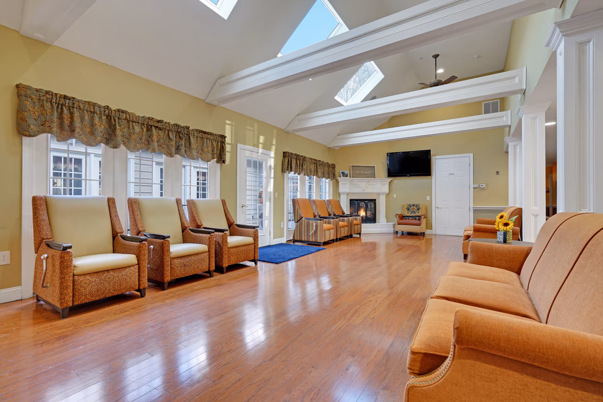 A spacious living room with wooden flooring, multiple upholstered armchairs and sofas arranged along the walls, a fireplace with a TV mounted above it, large windows with decorative valances, and skylights in the ceiling allowing natural light to fill the room.