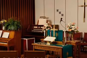 Interior of a chapel or worship area with a wooden piano, an organ, a pulpit with an open Bible, floral arrangements, candles, and a cross on the wall.