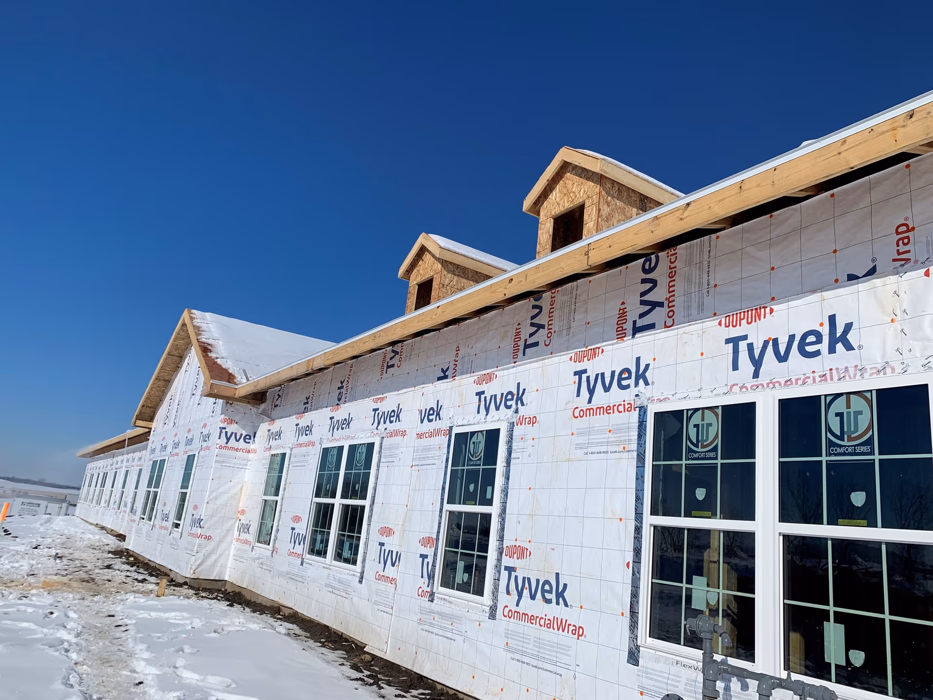 Exterior view of a senior living community building under construction, covered with Tyvek commercial wrap and featuring multiple windows. Snow is on the ground and the sky is clear and blue.