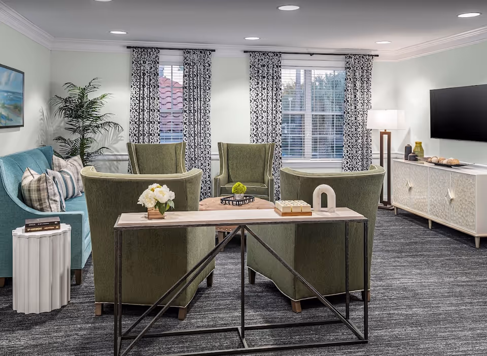 A cozy living room with four green armchairs arranged around a round ottoman, a teal sofa with patterned pillows, a white side table with books, a console table with decorative items, a floor lamp, and a wall-mounted flat-screen TV. The room has large windows with patterned curtains and a gray carpeted floor.
