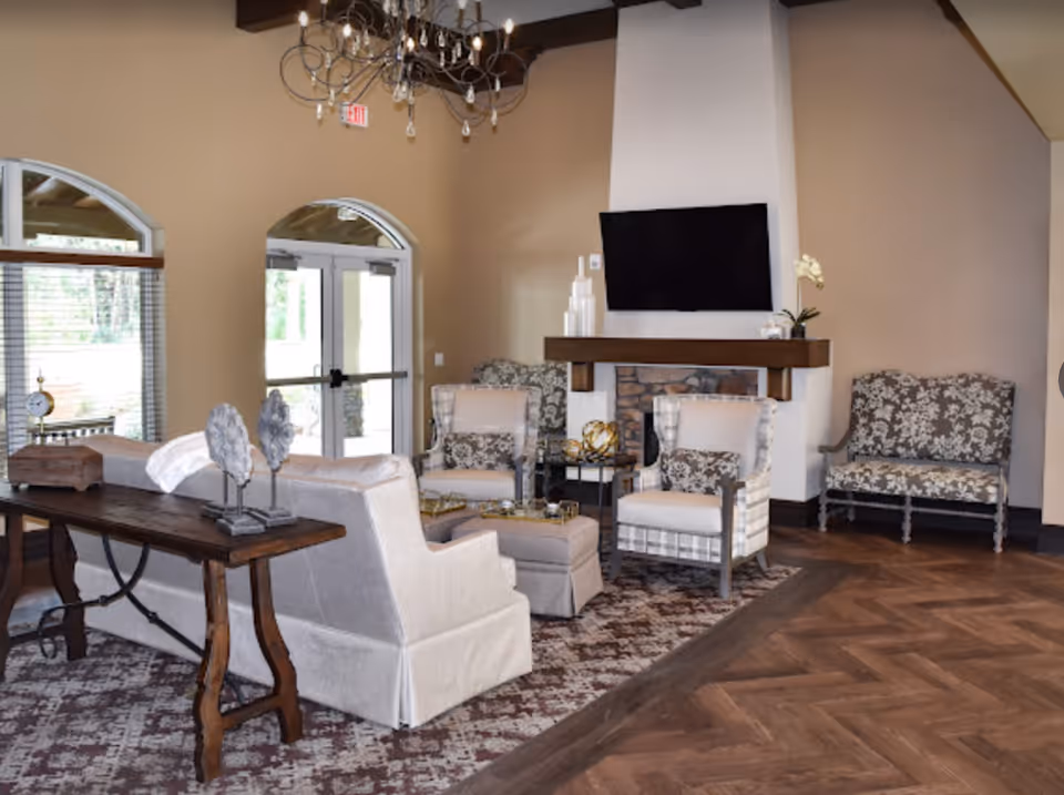A cozy senior living facility lounge area with beige walls, a wooden floor with a patterned rug, a white fireplace with a mounted flat-screen TV above it, and several upholstered chairs and a sofa arranged around a central ottoman. There is a wooden console table behind the sofa with decorative items, and large windows and glass doors letting in natural light.