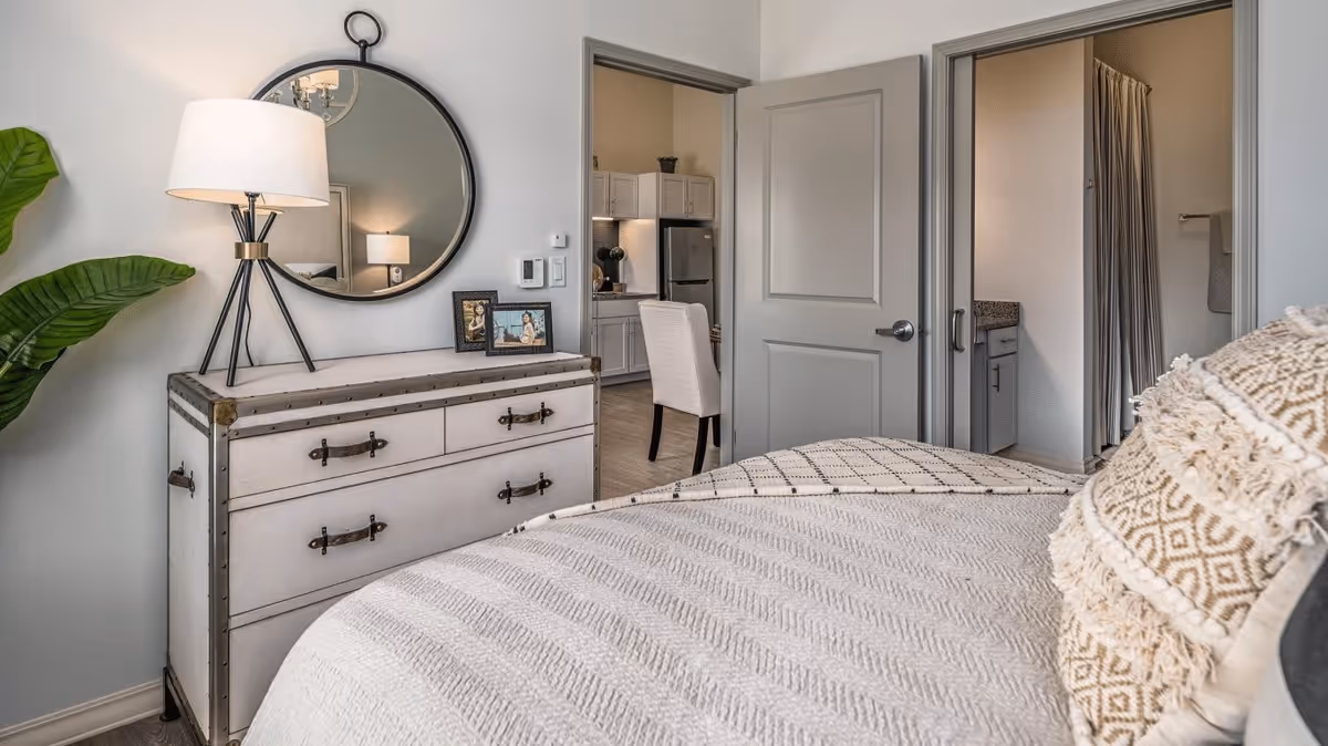 A cozy bedroom with a bed covered in a textured beige blanket and decorative pillows. To the left, there is a white dresser with metal accents, topped with a round mirror, a table lamp, and two framed photos. In the background, an open door reveals a kitchen area with white cabinets and a stainless steel refrigerator, while another door leads to a bathroom with a shower curtain and granite countertop.
