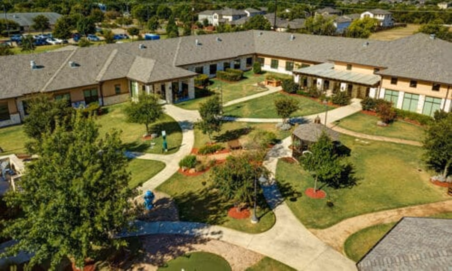 Aerial view of a senior living facility courtyard with green lawns, trees, walking paths, benches, and a gazebo surrounded by a single-story building with a gray roof.