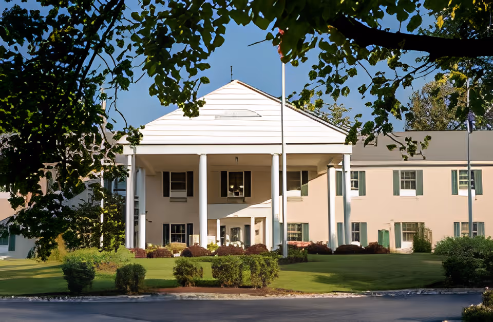 Two-story pale building with large white columns, a flagpole, and a manicured lawn framed by trees.