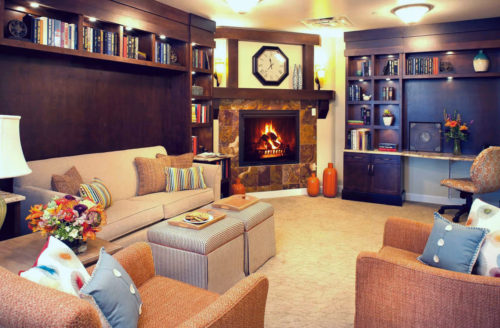 Cozy living room with a lit fireplace, beige sofa and armchairs, built-in bookshelves, ottomans, and decorative accents.