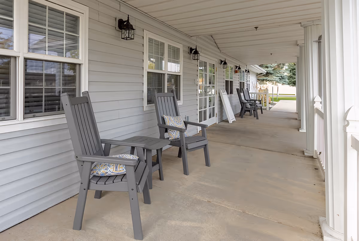Covered porch at a senior living facility with gray chairs and small tables along the building.
