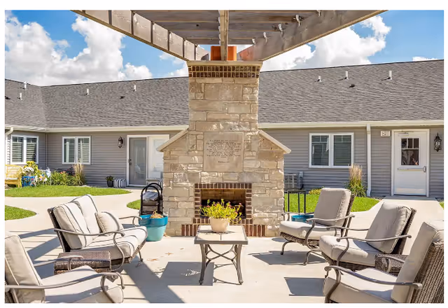Outdoor seating area with cushioned chairs arranged around a stone fireplace under a wooden pergola. The background shows a single-story building with multiple doors and windows, and a clear blue sky with some clouds.