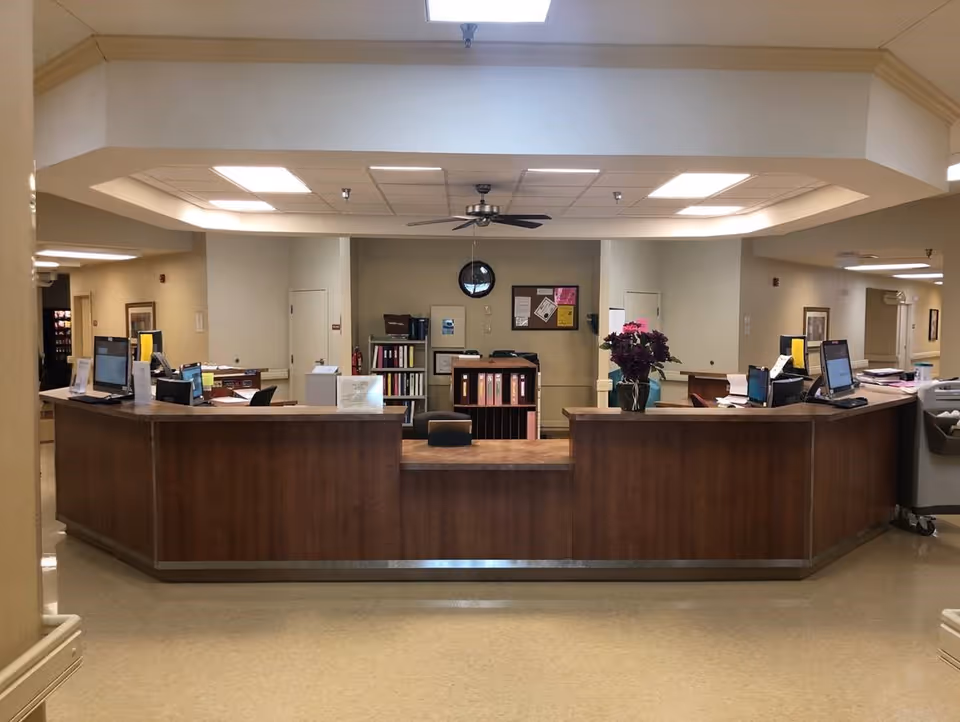 Reception desk area in a senior living facility with a wooden counter, computer monitors, office chairs, and shelves with binders and documents in the background. The ceiling has recessed lighting and a ceiling fan.