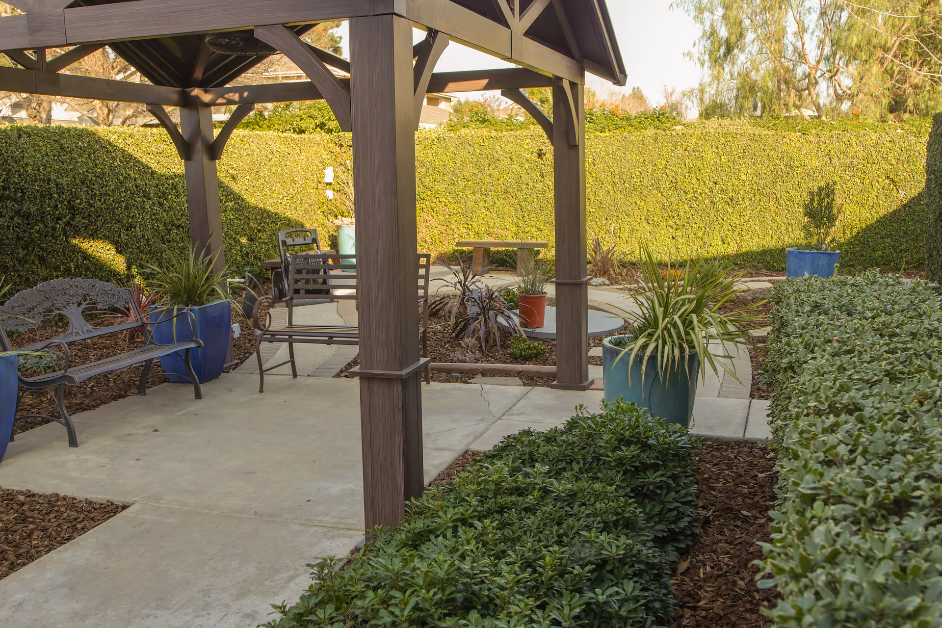Outdoor garden area with a wooden pergola, metal benches, potted plants, and neatly trimmed hedges surrounding a concrete pathway.