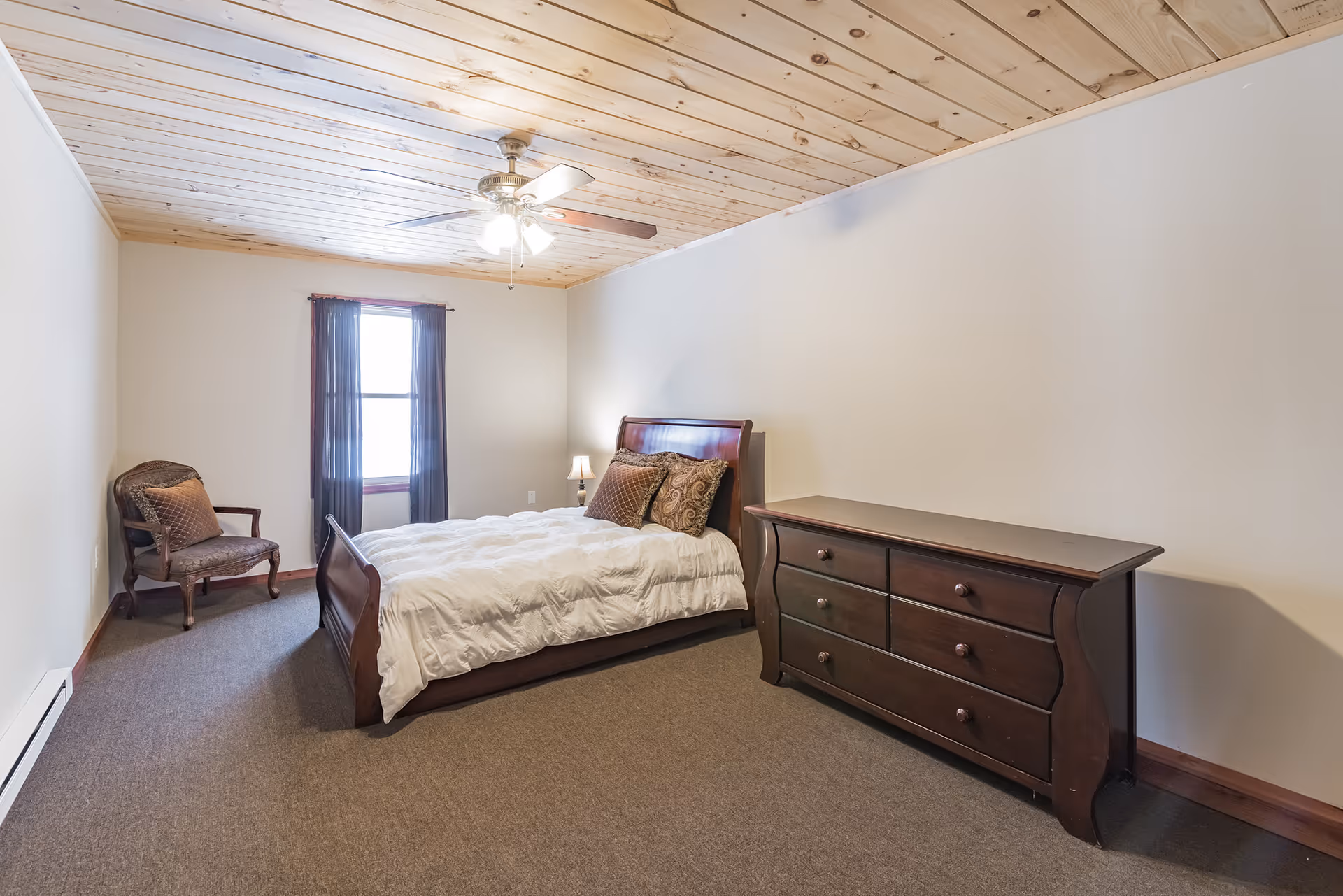 A bedroom with a wooden ceiling and beige walls featuring a bed with white bedding and decorative pillows, a dark wooden dresser, a small lamp on a nightstand, a window with dark curtains, and an upholstered chair in the corner.