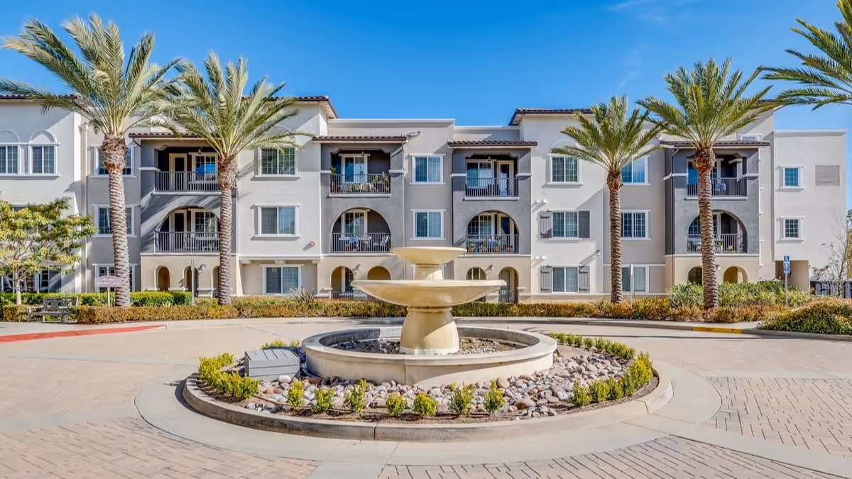 Exterior view of a three-story senior living facility building with balconies, palm trees, and a circular driveway featuring a central water fountain surrounded by landscaping.