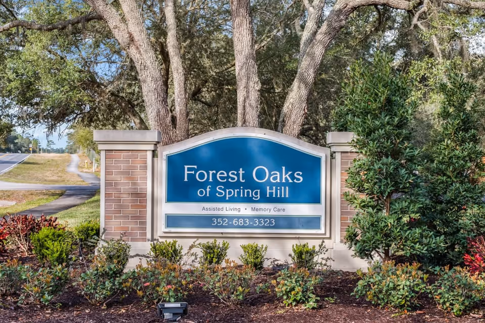 Outdoor entrance sign for Forest Oaks of Spring Hill, an assisted living and memory care facility, surrounded by trees and landscaping with a road visible to the left.