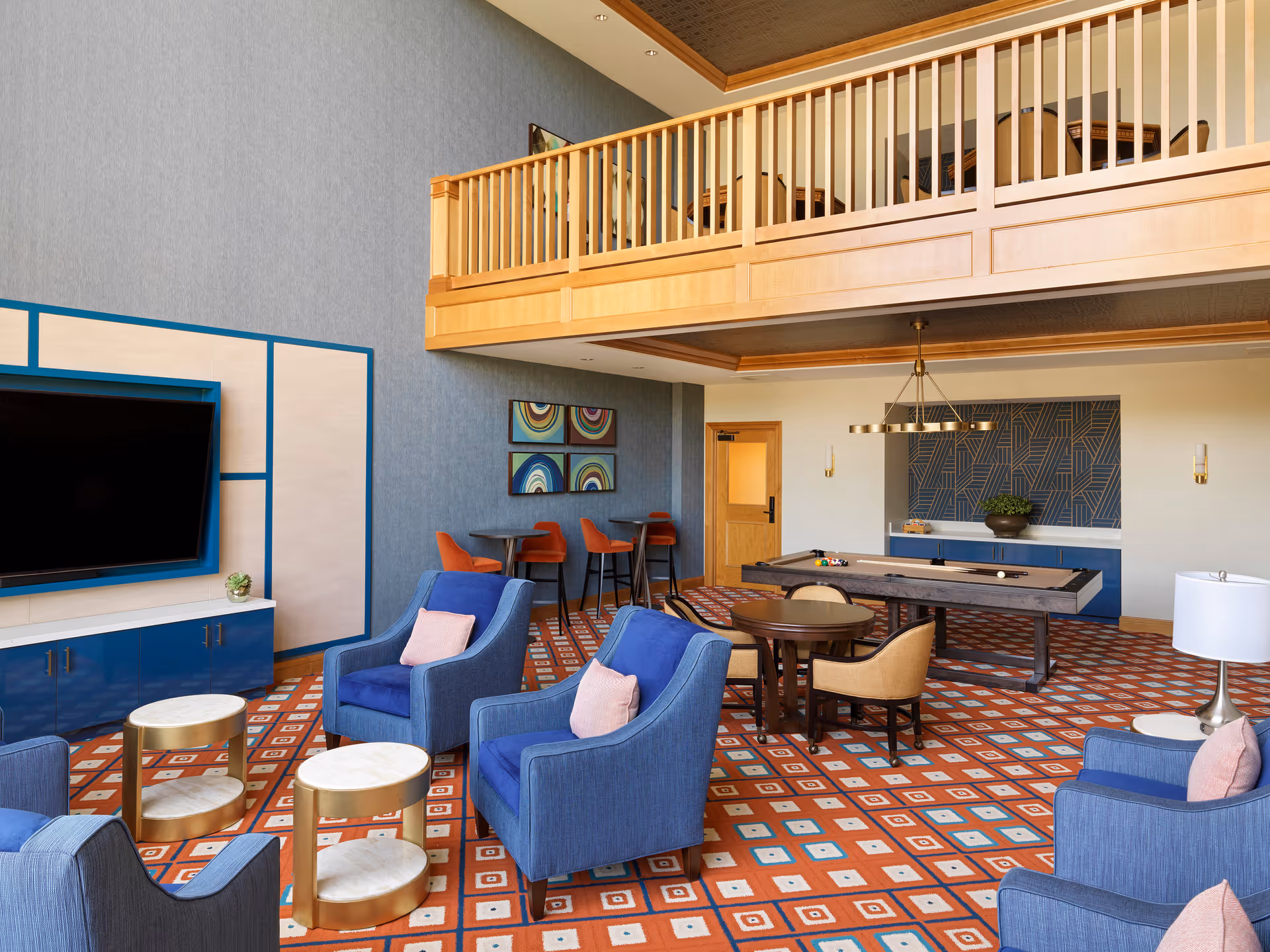 A spacious and stylish common area with blue armchairs and small round tables on a patterned orange carpet. There is a large flat-screen TV mounted on the wall, a pool table, and a small seating area with orange chairs and round tables. The room features a wooden balcony railing above and modern wall art.