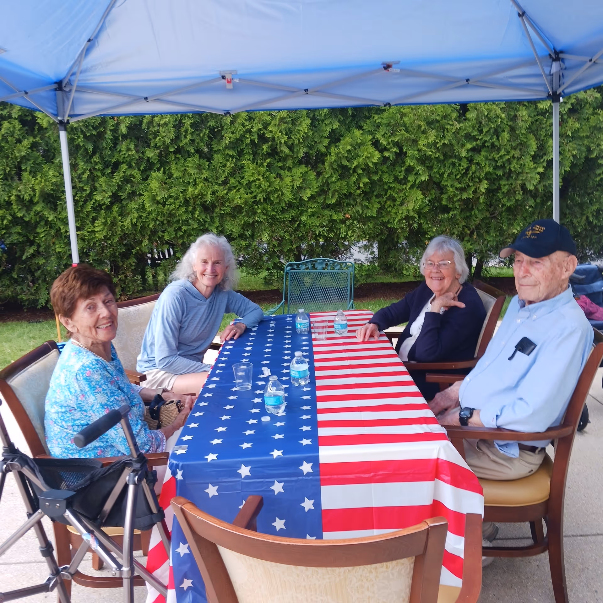 Four elderly people sitting around a table covered with an American flag tablecloth under a blue canopy tent outdoors, with green bushes in the background. There are water bottles and a plastic cup on the table.