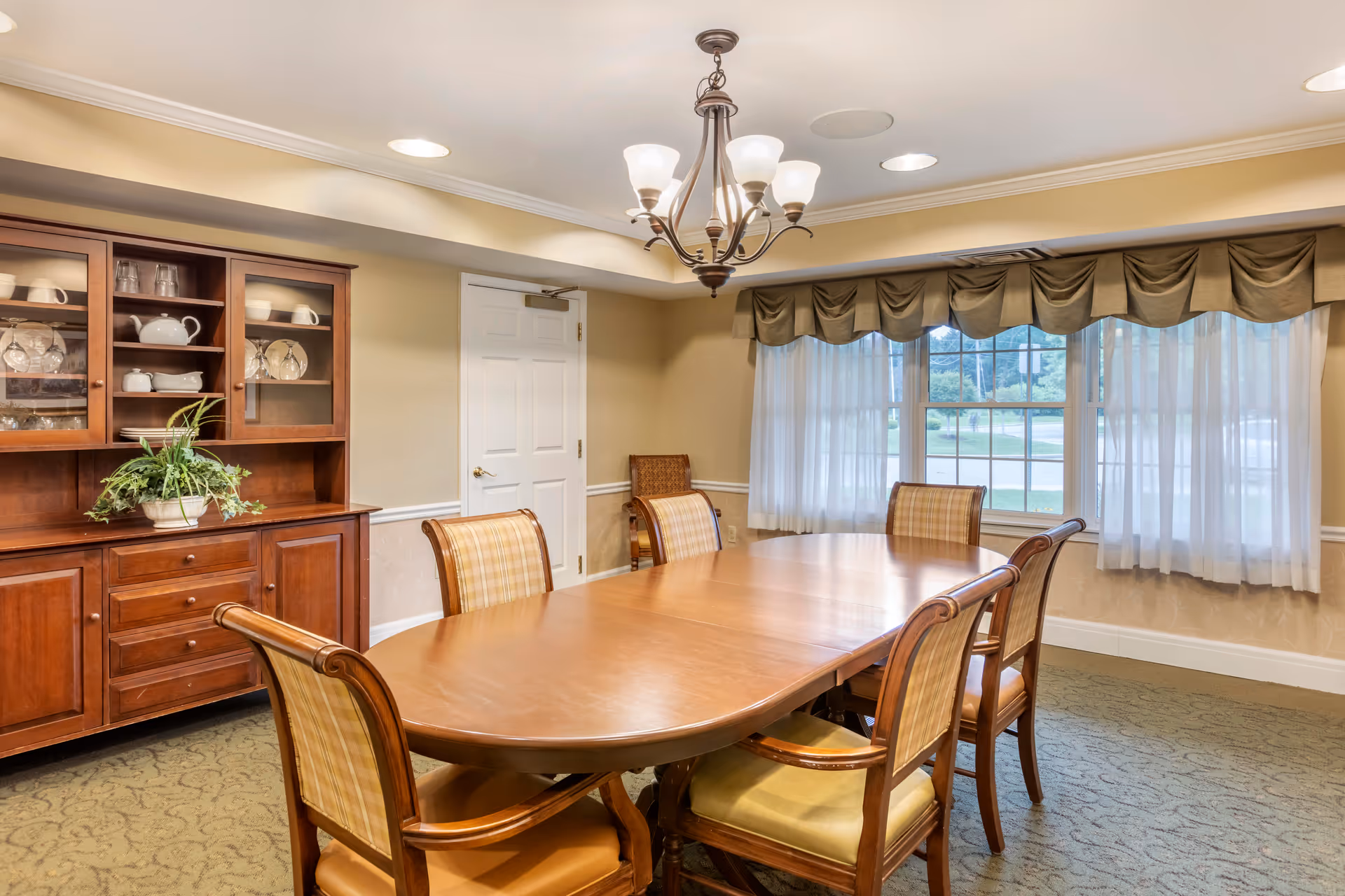 A well-lit dining room with a wooden dining table surrounded by six cushioned chairs. A wooden cabinet with glass doors displaying dishes and glassware is against one wall. A window with sheer white curtains and a valance lets in natural light. A chandelier with multiple lights hangs above the table.